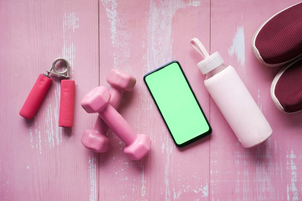 Dumbells, water bottle, hand grip gripper and mobile on a pink floor symbolising hydration and a healthy lifestyle